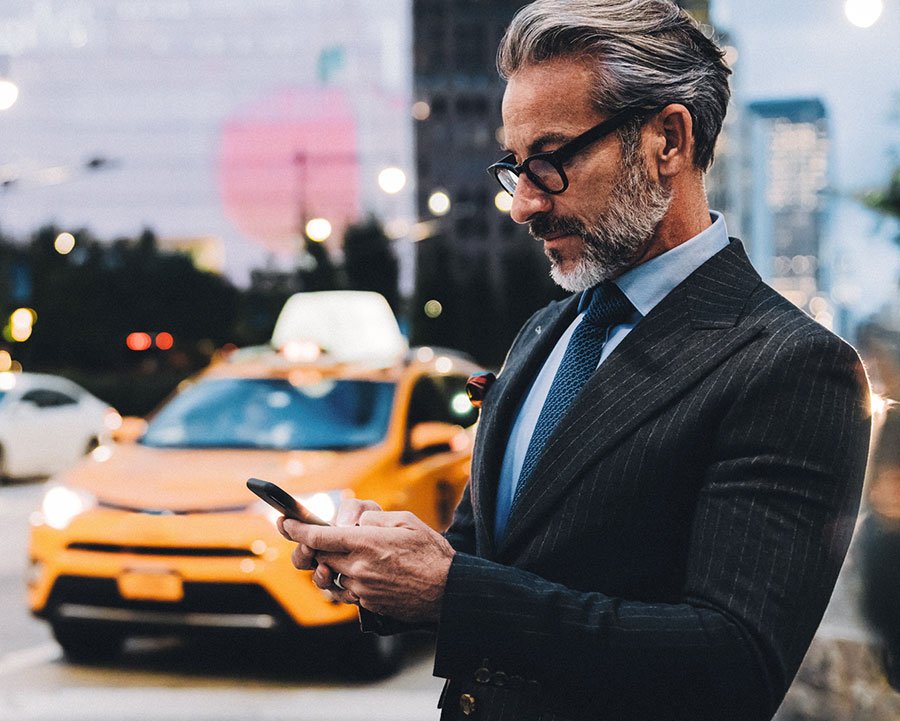 Middle age male financial professional with a beard and glasses standing along a busy city street with taxis driving by and texting his client about considering a hybrid QDIA for their retirement plan.